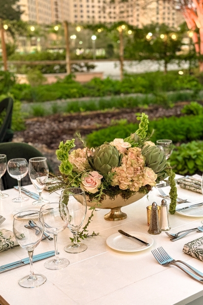 A beautiful centerpiece with pink flowers and artichokes sits on a set table amidst Emma's Creekside Farm at Rosen Shingle Creek for a post-wedding event.