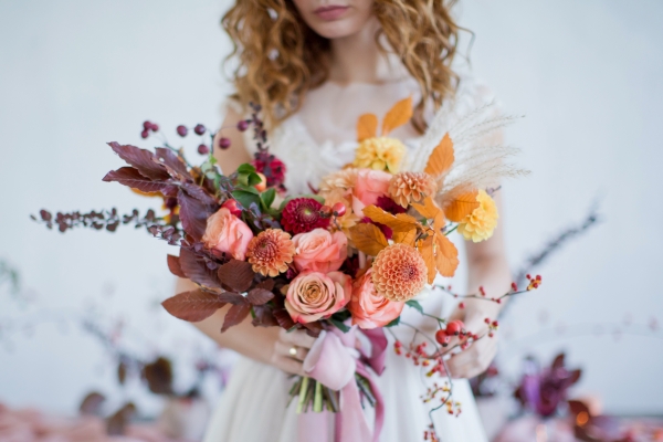 A bride in a white dress holds a bright and colorful fall bouquet with oranges, pinks, and other beautiful autumn hues.