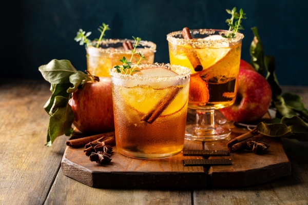 Three golden cocktails on a wooden board adorned with fresh apples, cinnamon sticks, and star anise.