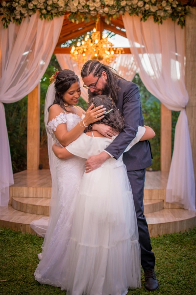 A bride and groom embrace a young girl in a white dress in front of an altar with drapery, flowers, and a glowing chandelier.