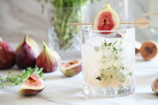 A cocktail garnished with sprigs of time and a sliced fig, on a table surrounded by figs.