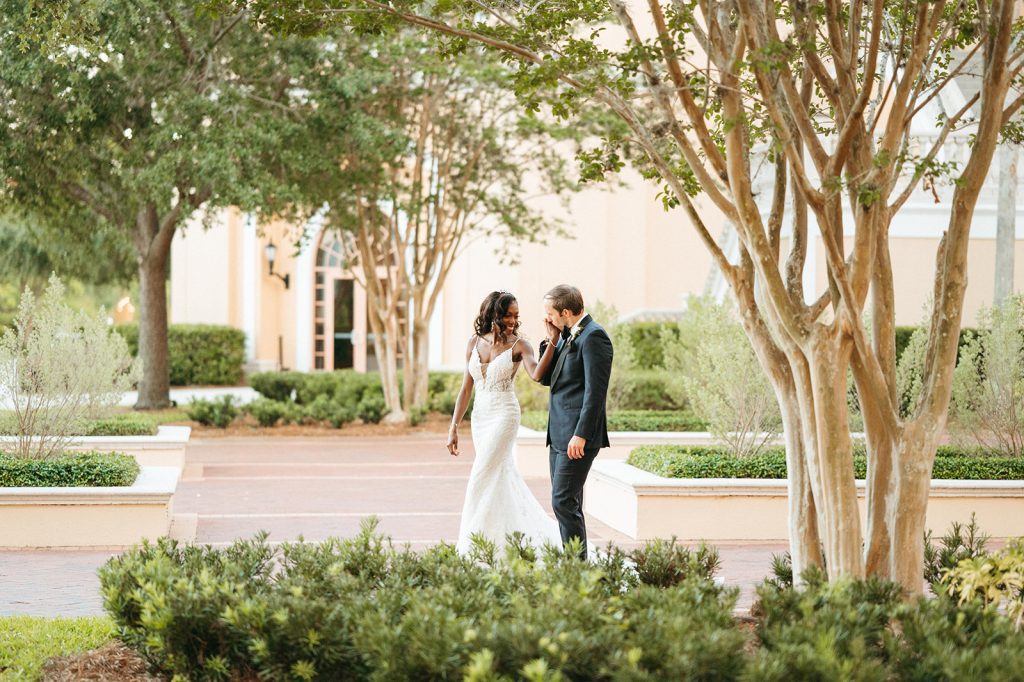 A bride and groom walk hand in hand along a courtyard lined in greenery and mature trees. 