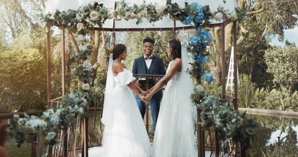 Two women in bridal gowns and veils hold hands beneath a decorated floral arch in front of a laughing man in a suit.