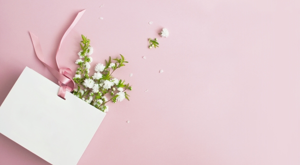 A white wedding welcome bag with pink ribbon lays on a pink background with white flowers pouring out of it.