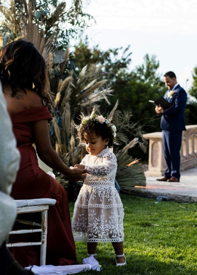 A girl in a lacy white dress holds hands with a seated woman in a red dress in an outdoor venue while an officiant in a blue suit stands out of focus in the distance. 