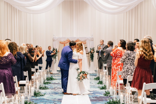 A bride and groom stop in the middle of the aisle to kiss between people standing on either side. 