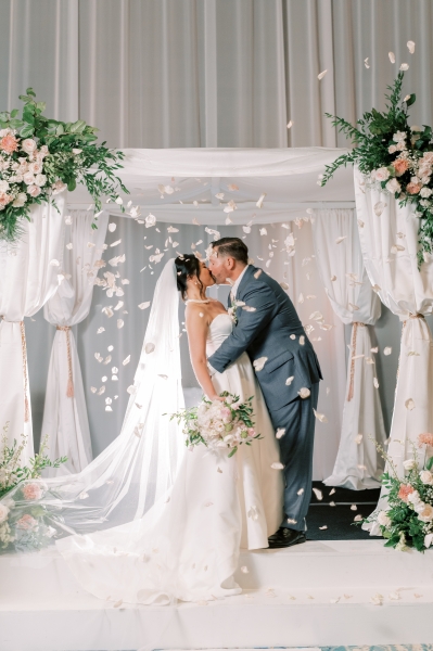 A bride and groom kiss under a canopy of white drapery with greenery and pink flowers, with floating flower petals flying through the air at Rosen Plaza during an indoor ceremony.