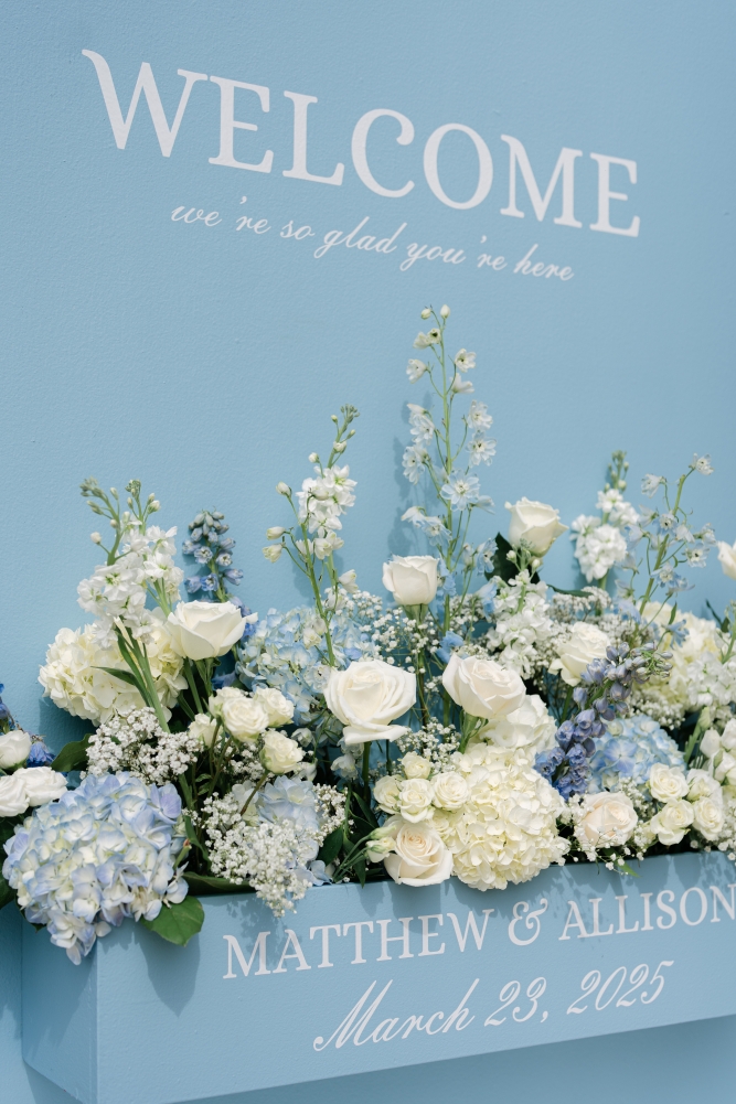 A long arrangement of blue and white hydrangeas, roses, and baby's breath in a light blue box that reads "welcome" and "Matthew & Allison, March 23, 2025".