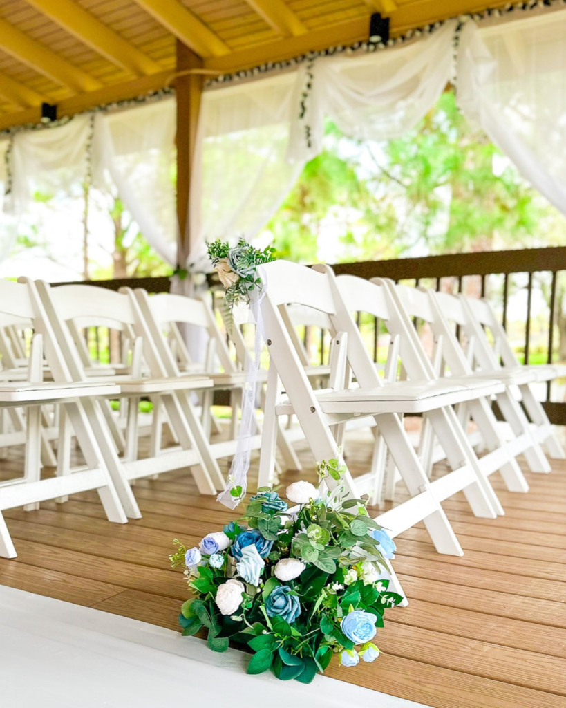 Rows of white folding chairs on a wooden patio with a close-up or a blue floral arrangement lining the aisle. 