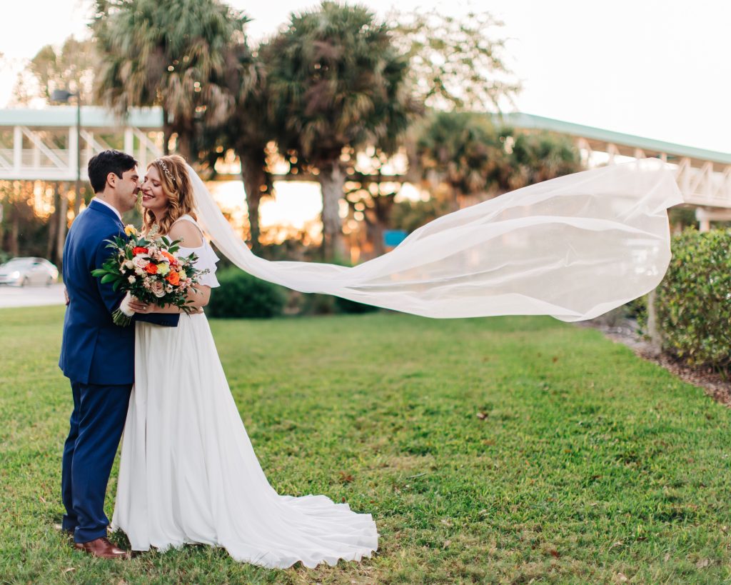 A bride and groom stand outside on a green lawn in front of palm trees embracing, the bride's veil blowing behind her.