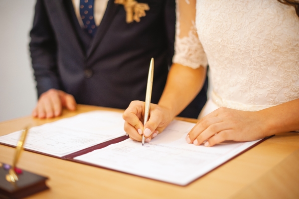 A woman in a white dress signs an official document standing next to a man in a suit.