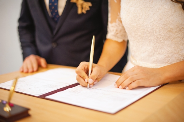 A woman in a white dress signs a formal document on a table beside a man in a dark suit. 