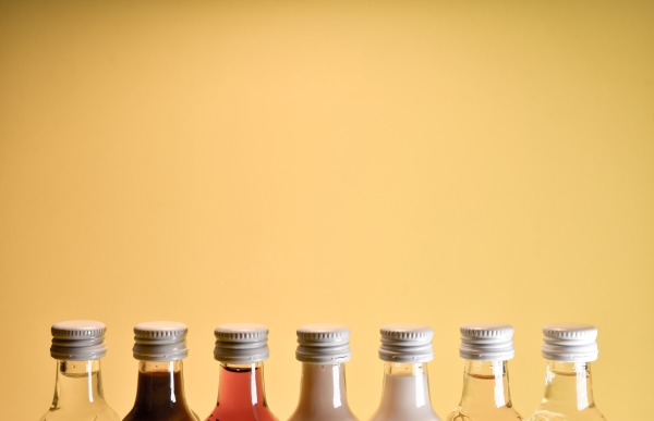 The tops of seven mini liquor bottles, each with a different colored liquid in them, against a yellow backdrop.