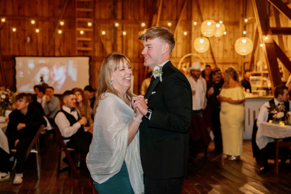 A woman in a shawl dances with a young man in a black suit in a barn lit with twinkle lights and full of onlookers.