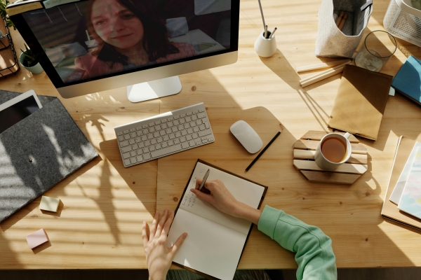 An overhead shot of a person taking notes in a journal in front of a computer screen surrounded by a mug of coffee, journals, pens, and other supplies.