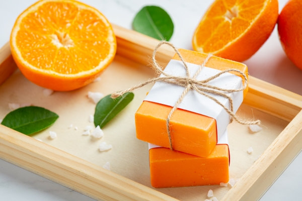 Bars of handmade orange soap tied with white paper and twine on a wooden tray with orange leaves and sliced oranges.