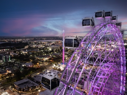 In the foreground, a large observation wheel glows bright purple while looking out at a lit up stretch of Orlando below at dusk. The Orlando Eye is a romantic thing to do on your Orlando honeymoon.