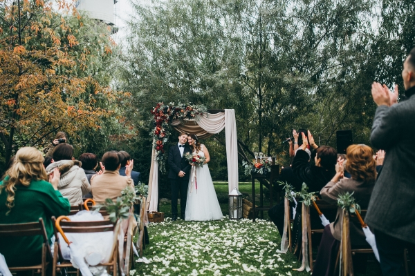 A bride and groom stand outdoors in front of an altar draped with fabric with friends and family seated in chairs facing them.