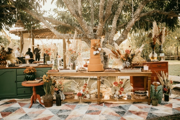 A four-tiered autumnal colored cake sits on a table surrounded by fall foliage and pampas grass and antique furniture at an outdoor event.