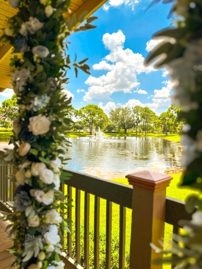 A view of a beautiful fountain surrounded by green grass, trees, and a bright blue sky, as seen through two pillars decorated with white florals and greenery.