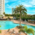 A large outdoor swimming pool with palm trees, lounge chairs, and tall buildings in the distance.