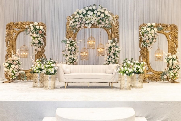 Three ornate golden arches decorated in white and light pink florals with suspended hanging birdcages sit in front of a wall covered in white drapery and surrounding a white sofa. 