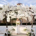 A bride in a white gown holding a bouquet walks beneath an archway of branches and white flowers along an aisle between empty white chairs on an outdoor patio.