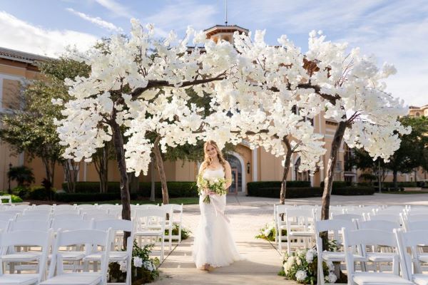 A bride in a white gown holding a bouquet walks beneath an archway of branches and white flowers along an aisle between empty white chairs on an outdoor patio.