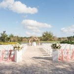 A large cobblestone patio set up with white chairs and pink drapery, columns with ornate floral displays, and in the distance, a geometric altar adorned with drapery and flowers. In the background is a lake, trees, and an open blue sky. Whether or not you decide to have kids at your wedding, Rosen Weddings can make your dreams come true.