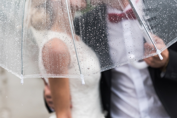 The torsos of a bride and groom beneath a transparent umbrella covered in raindrops.