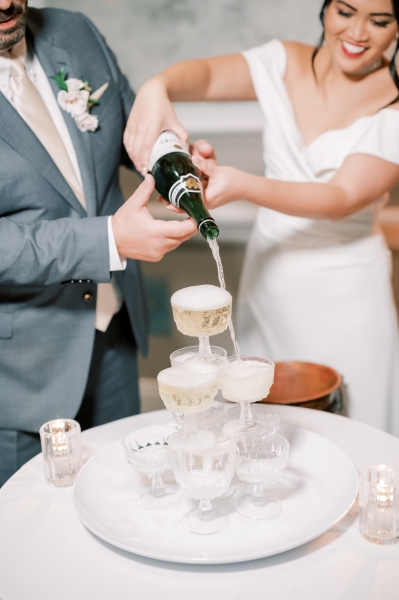 A smiling bride and groom pour a bottle of Champagne onto a tower of Champagne glasses during their wedding at Rosen Plaza.