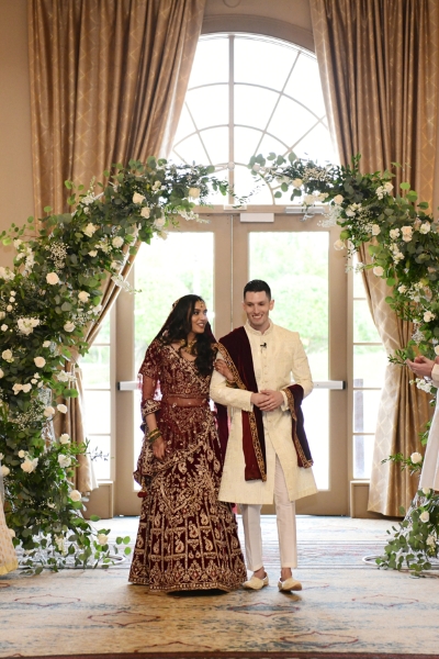 A bride and groom in traditional Indian clothing under a curved archway with greenery during an indoor wedding ceremony at Rosen Shingle Creek.