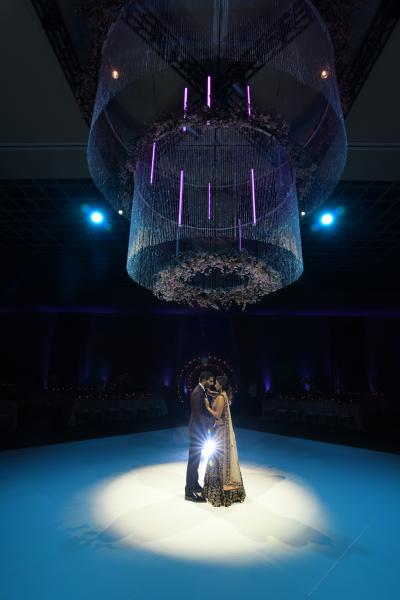 A couple in traditional Indian wedding attire dance in a dark room beneath a magnificent tiered modern chandelier. 