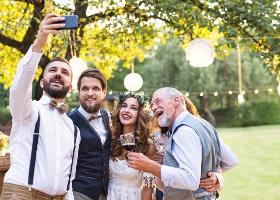 A bride and groom pose for a selfie with guests beneath a large tree decorated in lights.
