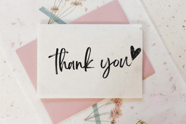 A white card that says "thank you" resting on a pink envelope and a paper background with pressed pink flowers.