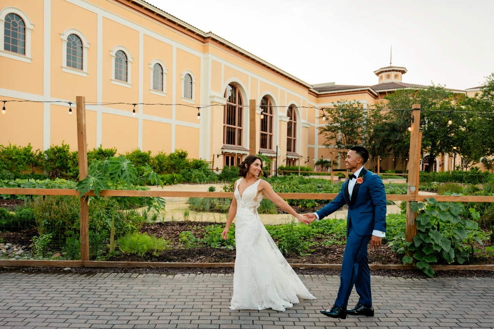 A bride and groom walk hand-in-hand in front of a garden and the stately Rosen Shingle Creek hotel. 