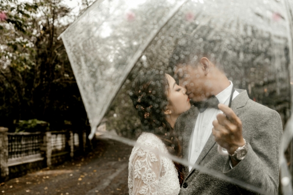 A woman in a white lacy dress kisses a man in a gray suit with a white shirt and black bowtie outdoors beneath a clear umbrella. 