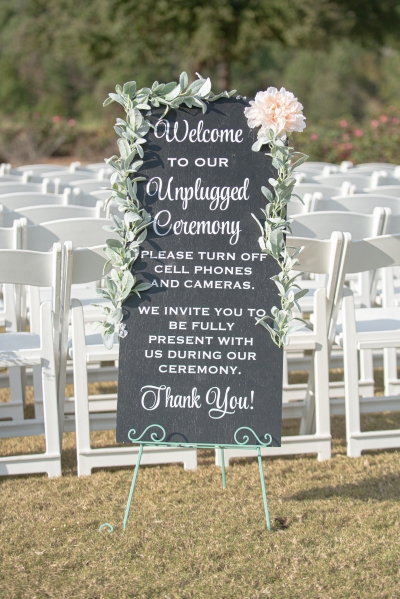 A chalkboard sign welcoming guests to an unplugged ceremony set in front of rows of white chairs in an outdoor setting.