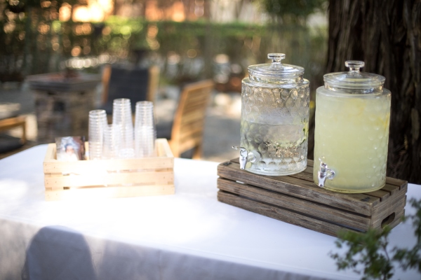 An outdoor table with a white tablecloth and a wooden crate displaying two glass carafes of water and lemonade.