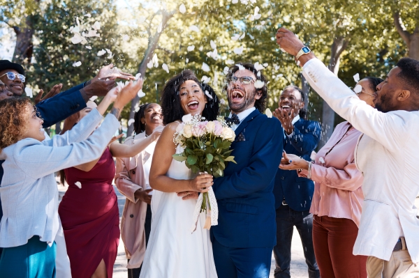 A bride and groom stand in the middle of a crowd of people throwing flower petals and cheering.