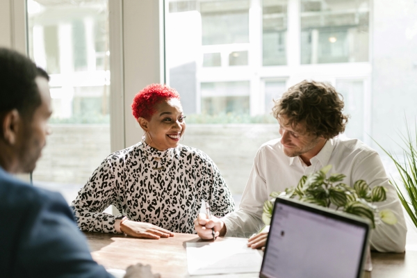 A woman with short red hair in an animal-print shirt sits at a table next to a man in a white button-down who is signing a document. A man in a suit with a computer sits across the table from them.