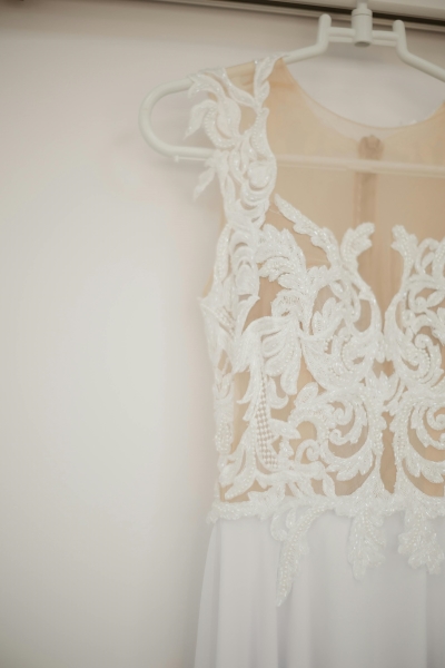 A close-up of a lacy wedding dress hanging against a white backdrop. Wedding dress preservation is a common unexpected expense. 