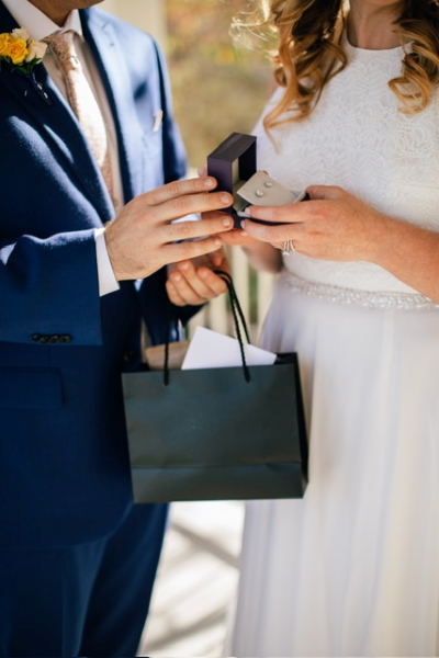 A man in a blue suit holding a black gift bag hands a woman in a white dress a box with diamond earrings.   