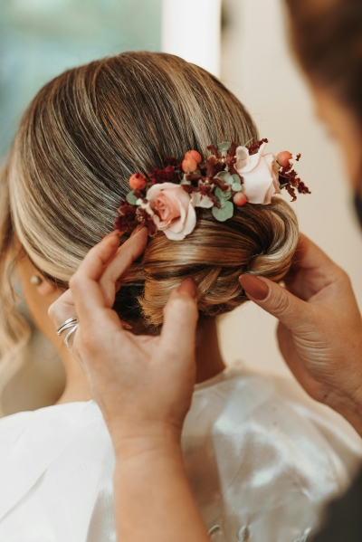 A woman’s hands with sienna-colored nails adjust the back of a woman’s hair in a smooth, formal bun with pink roses and maroon floral accents.  