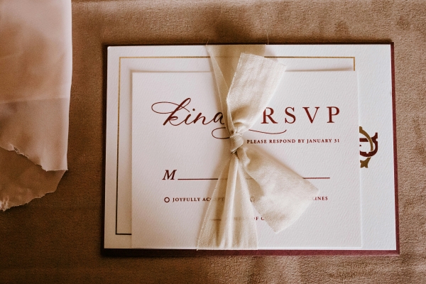 A white RSVP card with dark red ink tied to a white invitation with a beige gauze ribbon against a suede background. 