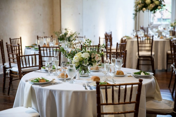 A circular table with white linens, a white floral centerpiece, and brown Chiavari chairs set with salads, dinner rolls, and water glasses. 