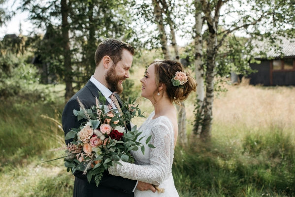 A woman in a lacy long sleeved white dress embraces a man in a dark suit, holding an autumnal bouquet in a grassy outdoor setting.