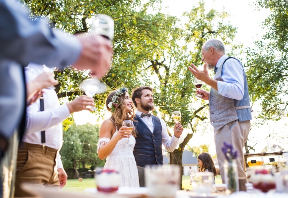 A bride and groom holding wine glasses smile at an older man in a vest speaking while at an outdoor celebration.