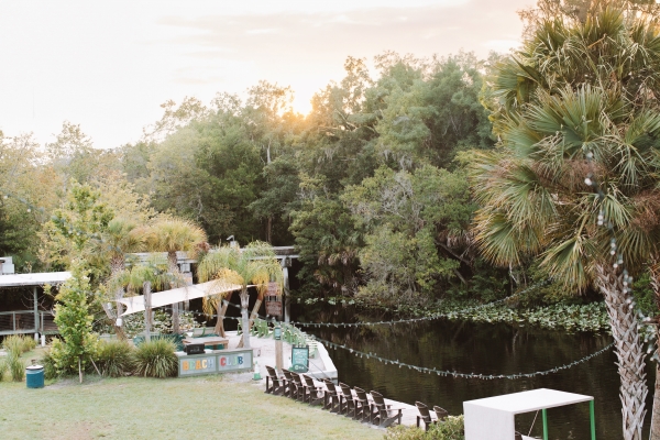 A river surrounded by lush palm trees and greenery with a dock of chairs and small buildings on one side.