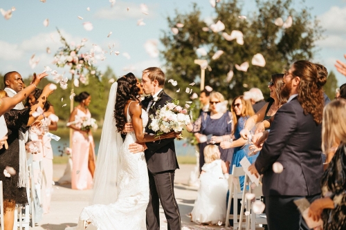 A couple kiss outdoors after walking down the aisle, surrounded by people throwing flower petals.